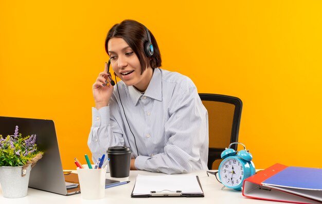 impressed-young-call-center-girl-wearing-headset-sitting-desk-putting-hand-headset-isolated-orange_141793-77509